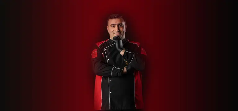 Kırmızı fonda, siyah-kırmızı aşçı ceketiyle gülümseyen Mahsun Usta